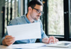 Serious pensive thoughtful focused young casual business accountant bookkeeper in office looking at and working with laptop and income tax return papers and documents