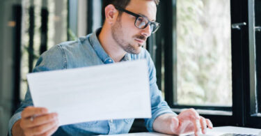 Serious pensive thoughtful focused young casual business accountant bookkeeper in office looking at and working with laptop and income tax return papers and documents